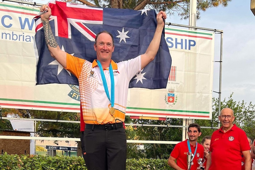 A man in an athletics shirt holds an Australian flag aloft while standing on a podium 