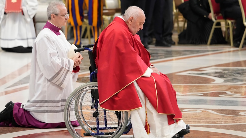 An elderly man in a red robe sits bowed in a wheelchair on a rich-looking floor.