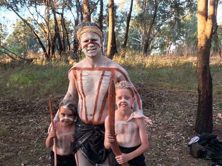 Indigenous man Joe Williams with his two young sons painted with ochre and holding boomerangs.
