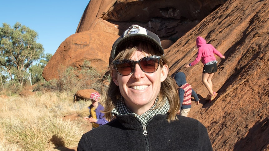 Megan Holbeck smiling in front of a large rock, kids playing in the background.