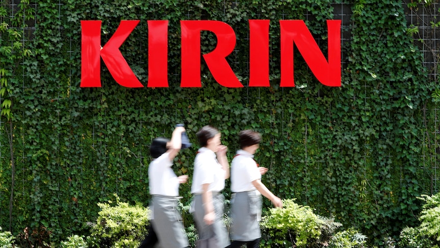 Employees are blurred as they walk past a big red Kirin sign with green ivy on the wall around it.