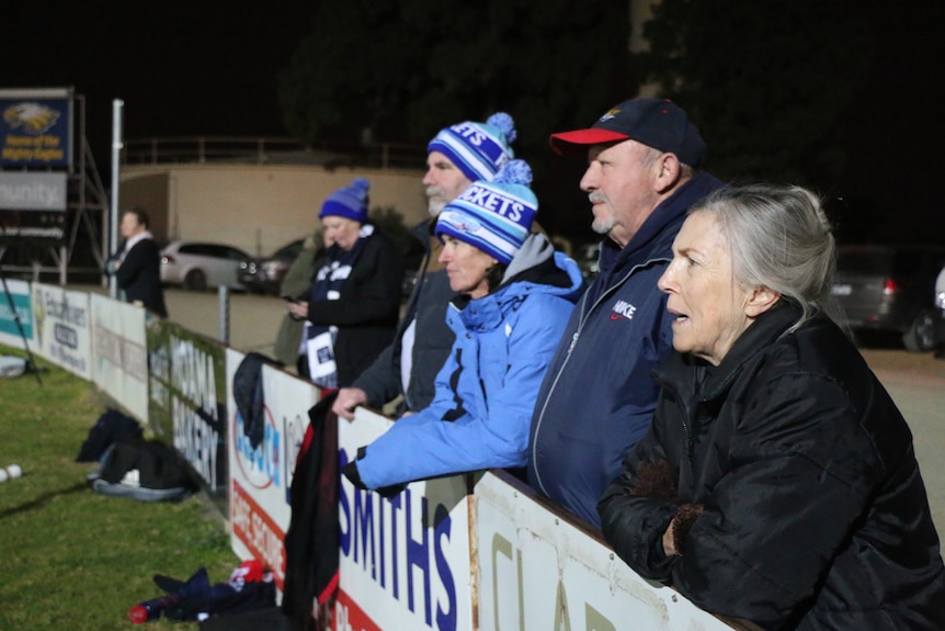A row of parents leaning over the fence at a footy oval watching training, three are wearing blue Rockets beanies.