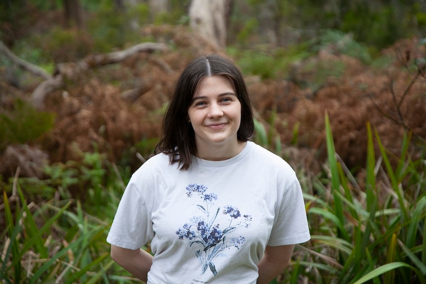 A young woman in a white t-shirt in front of a bush backdrop smiles