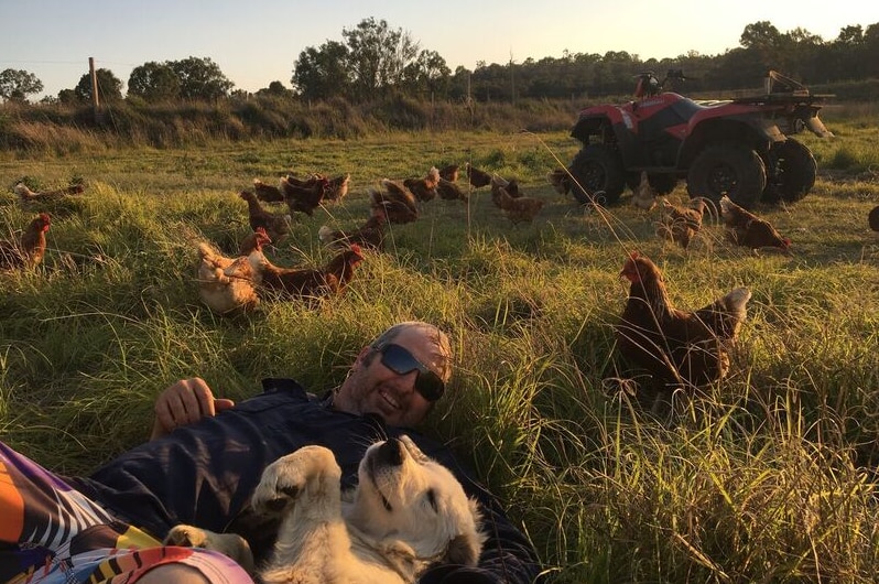 Dean Mayne lying in a grassy paddock with his dog and chickens around him and a quad bike in the background.
