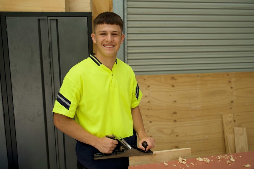 a young man in a high vis shirt shaving wood at a table and smiling