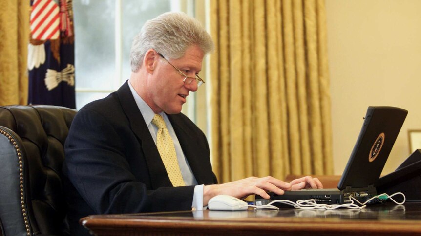 In the Oval Office, former US president Bill Clinton looks down at an early laptop while wearing glasses.