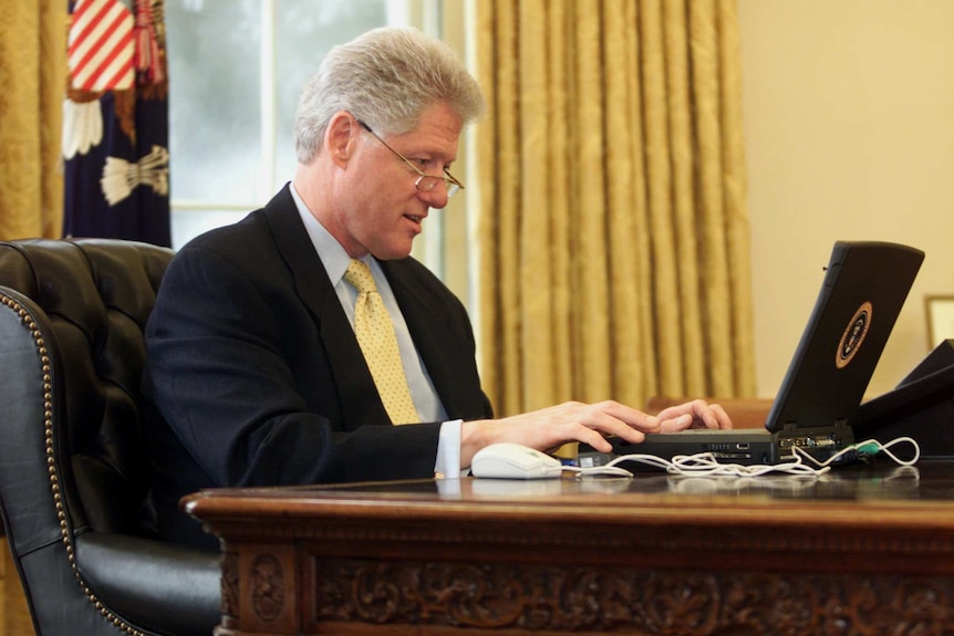 In the Oval Office, former US president Bill Clinton looks down at an early laptop while wearing glasses.