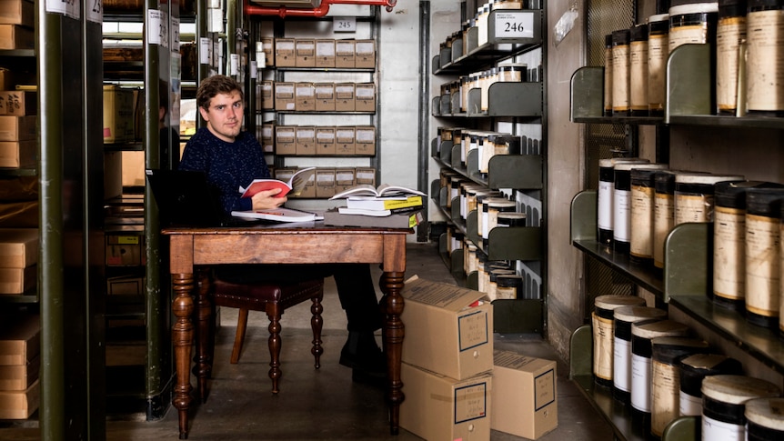 Man sits at desk with books, surrounded by archives.