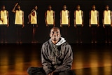 Young man in black pants and hoodie sits on gallery floor with video work featuring ten figures of him standing is behind.