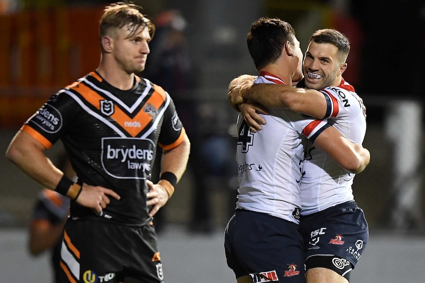 Two Sydney Roosters NRL players embrace as they celebrate a try, as a Wests Tigers opponent stands in the foreground.