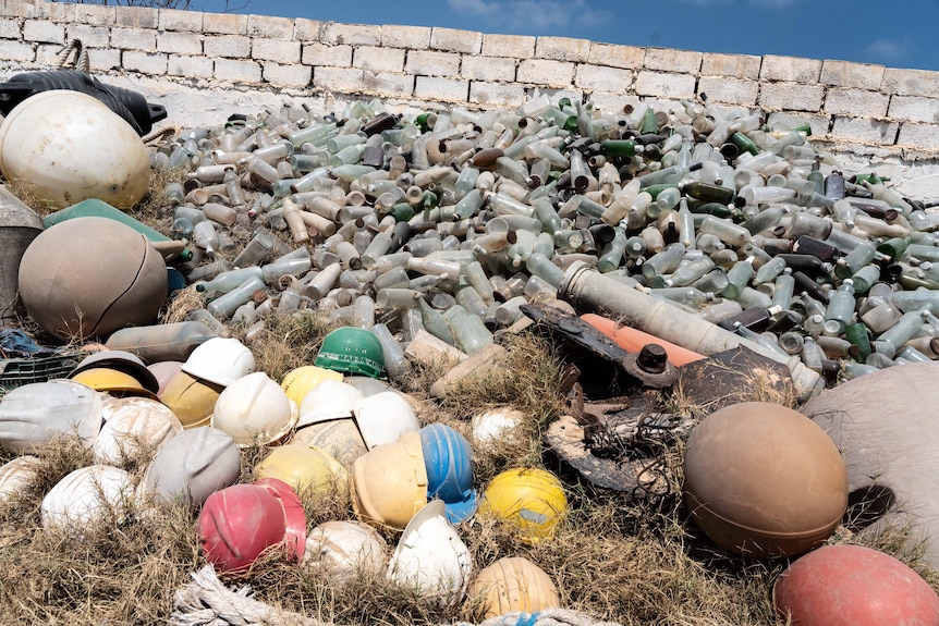A collection of ocean buoys in all sorts of colours sit on the grass stacked up against a brick wall by the ocean