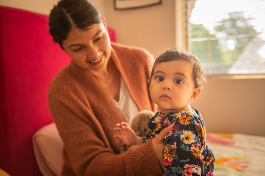 A baby girl looks up at the camera, with her smiling mother in the background.