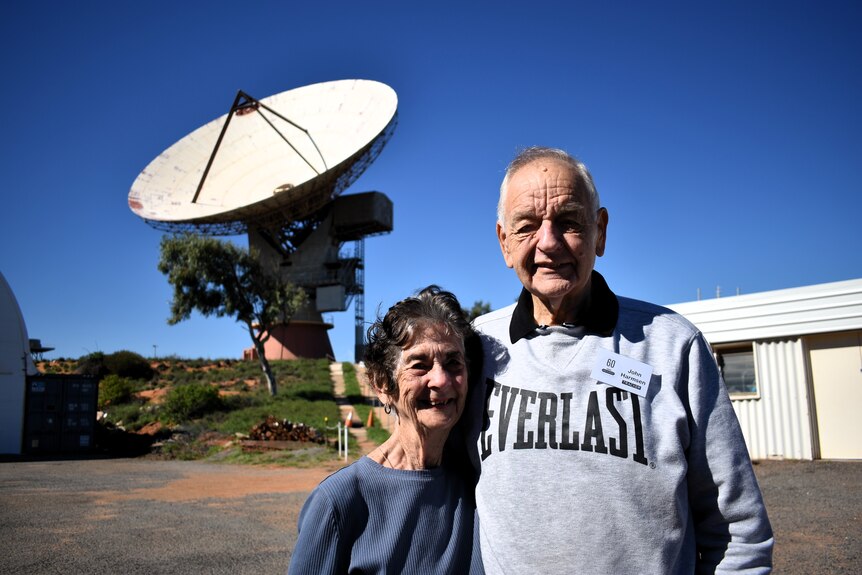 A man and woman in their eighties smile into camera, with a huge satellite dish pointed skywards behind them.