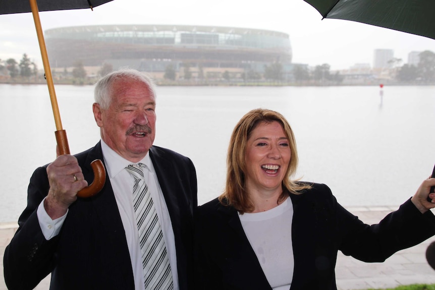 WA Ministers Mick Murray and Rita Saffioti stand holding umbrellas in front of the Swan River and Perth Stadium.