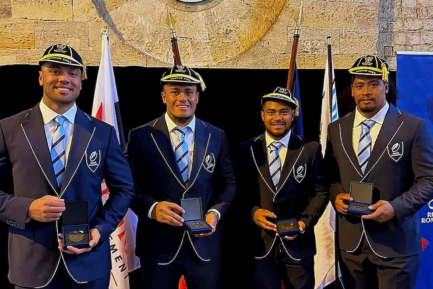 Four tongan men stand in blue suits, with rugby test caps on their heads, and are holding medals.