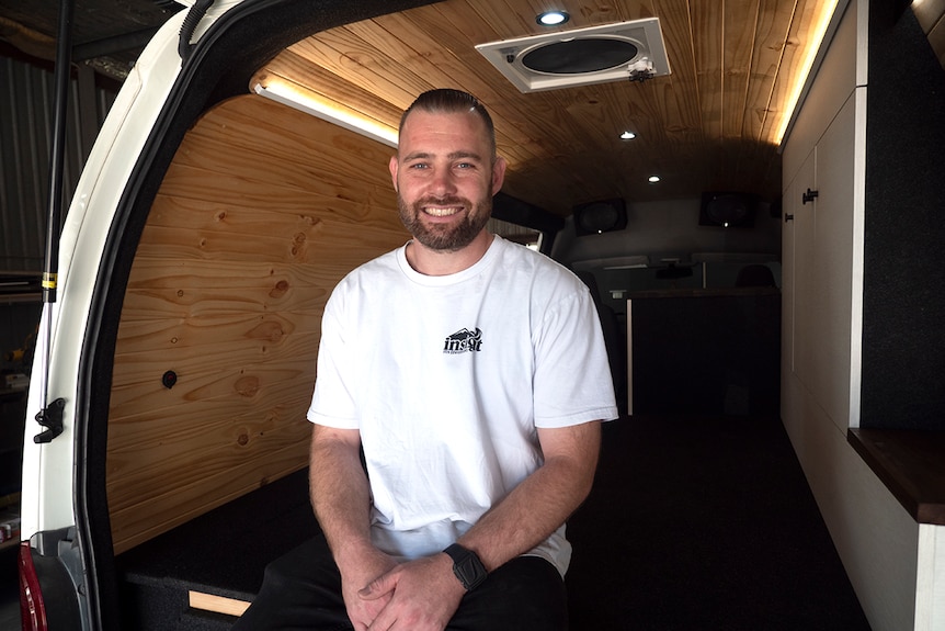 A man sits inside a van that has been fitted out for sleeping cooking and travelling.
