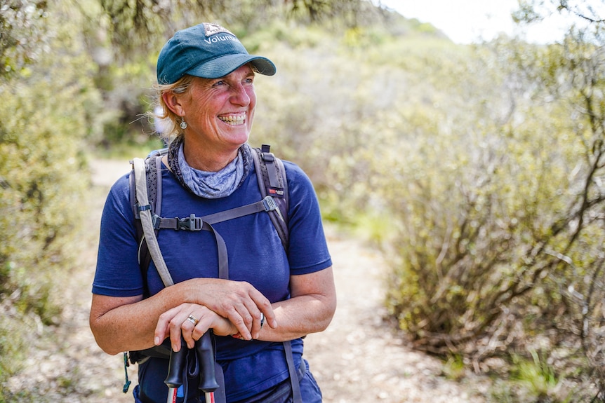 A woman wearing a blue shirt, cap and backpack stands on a leafy track.