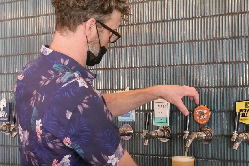 A man pours a beer from a line of beer taps on a tiled wall.