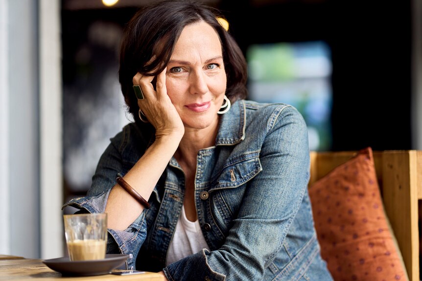 A middle-aged white woman with brown hair gazes at the camera, her chin in her hand, a half-finished latte in the foreground