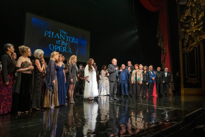 A theatre cast lined up on a satge with phantom of the opera written on a screen behind them