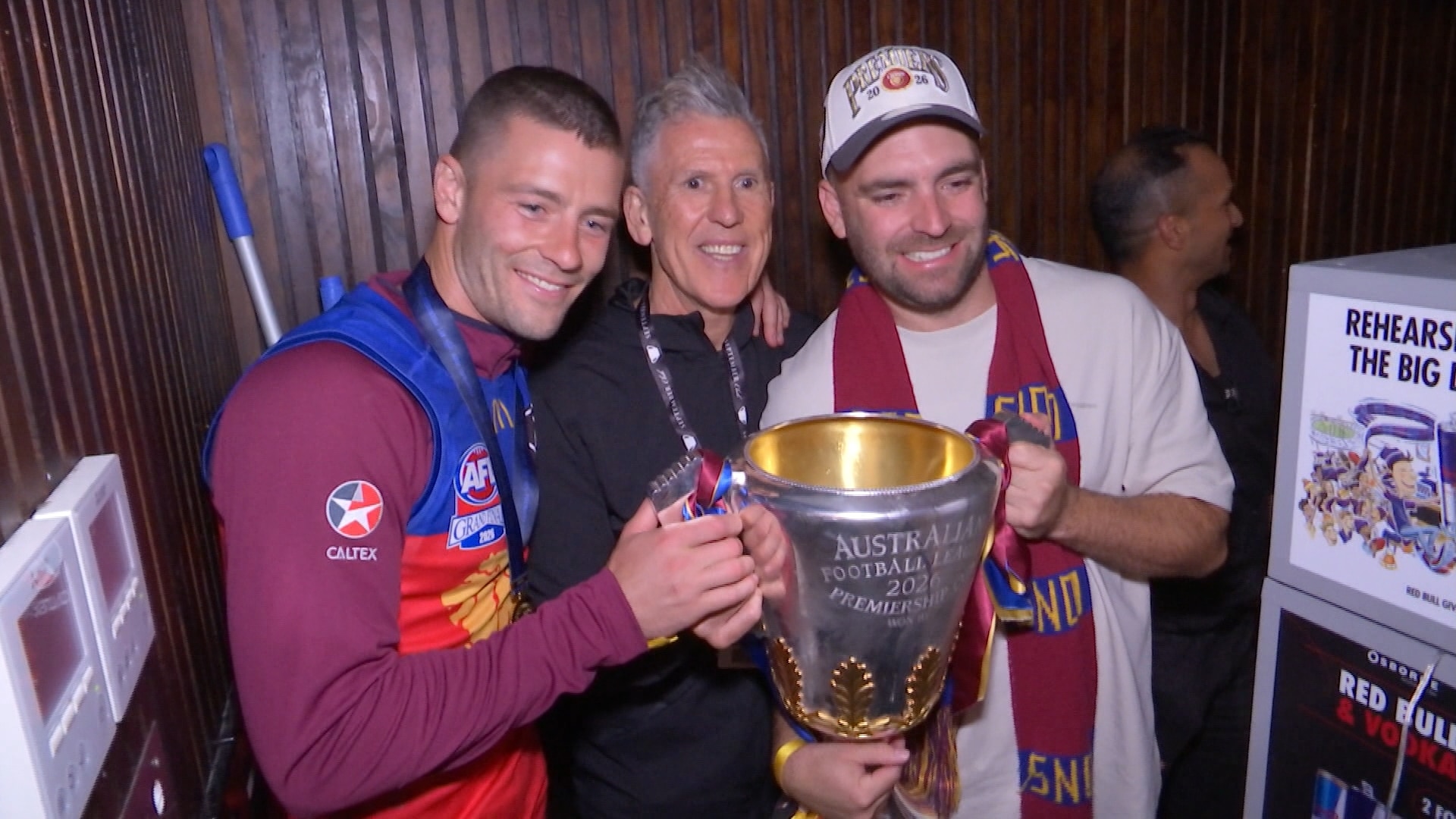 Three men, some dressed in Brisbane Lions gear, stand arm in arm holding a big silver cup.
