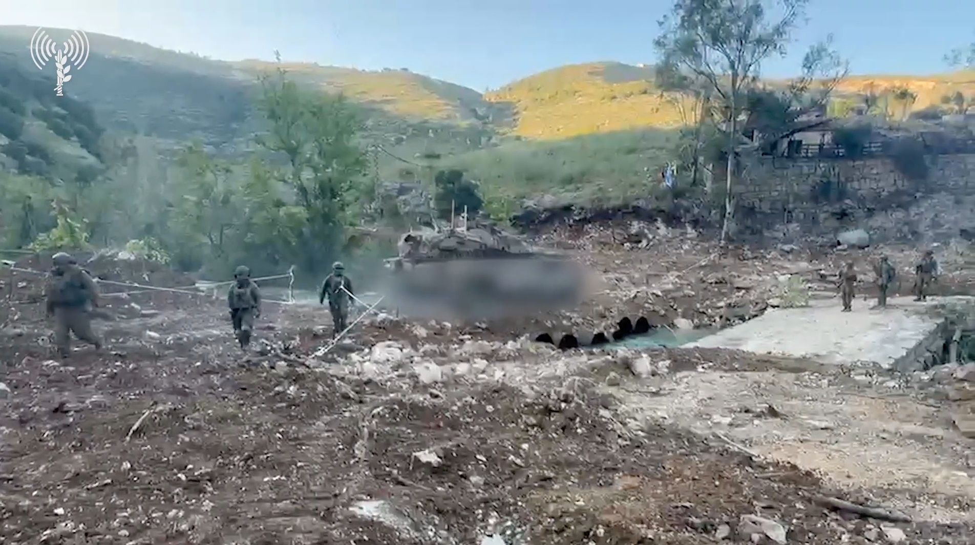 Los soldados se paran y caminan detrás de un tanque borroso. En primer plano, el terreno es aplanado, con colinas al fondo.