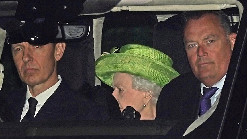 The Queen in a green hat sits in the back of a car with two men in the front seats.