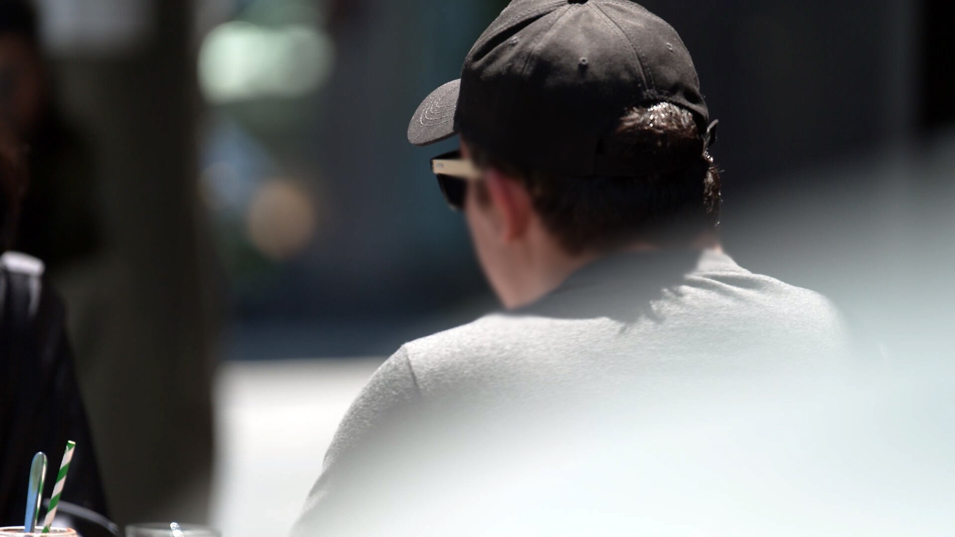 The back of the head of a man wearing a cap and glasses, sitting outside. His face cannot be seen.