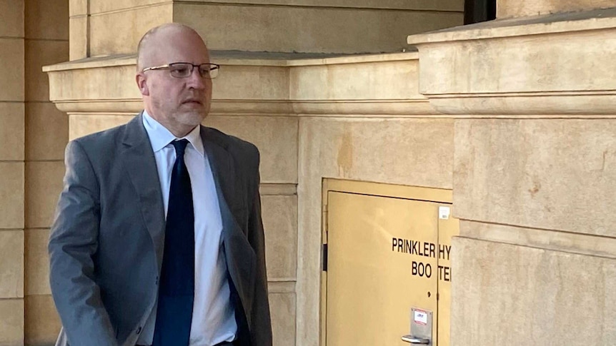 A man in spectacles and a grey suit walks along the footpath beside courts in Adelaide.