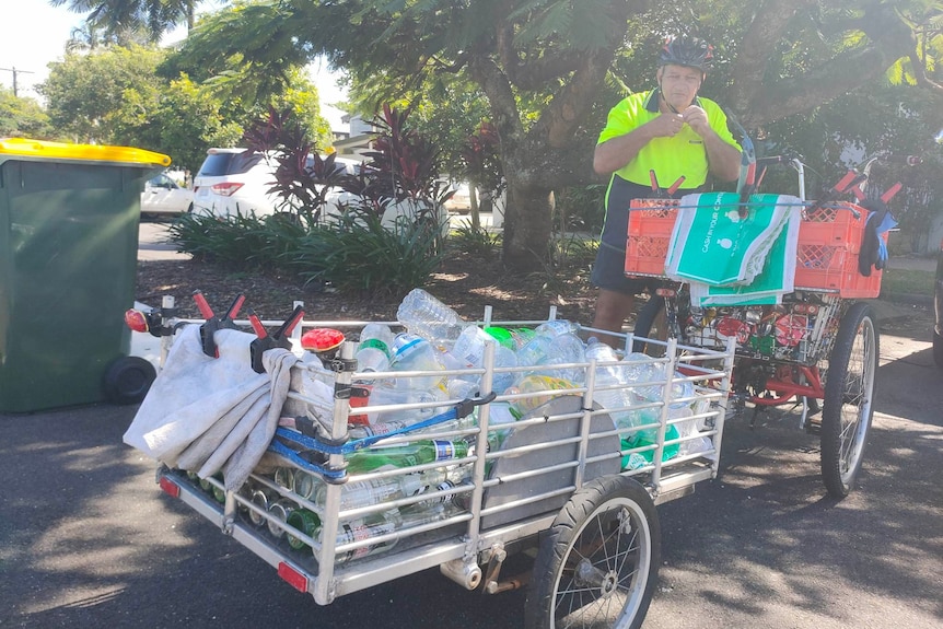 A trailer full of used bottles and cans is linked to a red adult tricycle while a man in a yellow shirt clips on his helmet