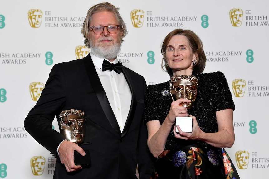 Two writers stand holding BAFTA trophies.