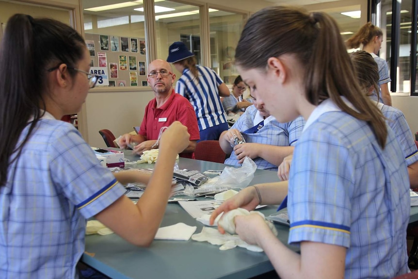 A male teacher sits in a room of girls making birthing kits