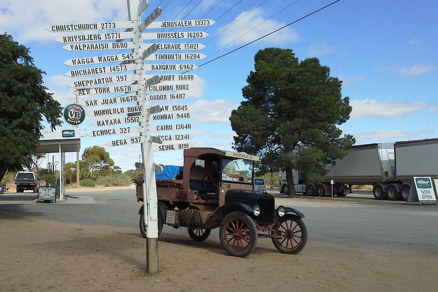 A vintage car parked beside a sign that shows the distance to cities around the world