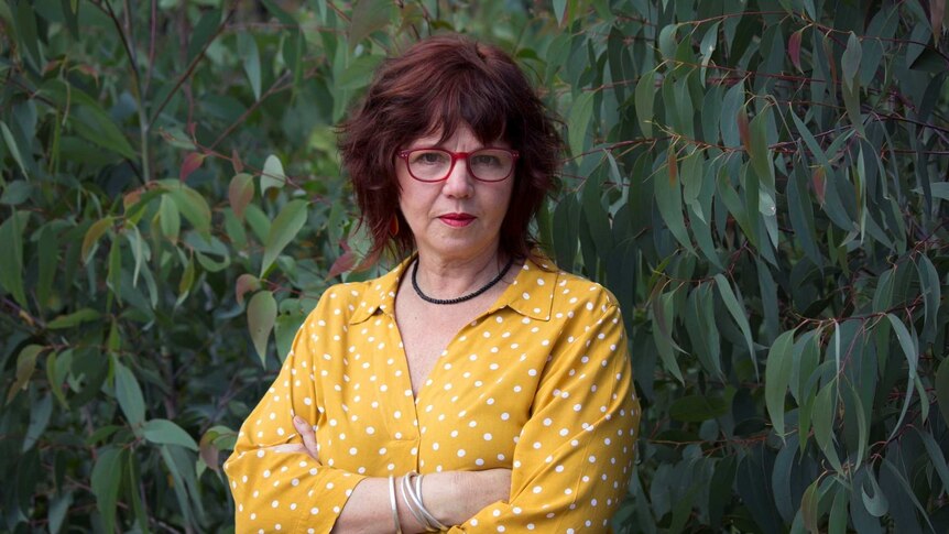 Jo Dodds stands of a gum tree wearing red glasses and a yellow shirt looking directly into the camera.