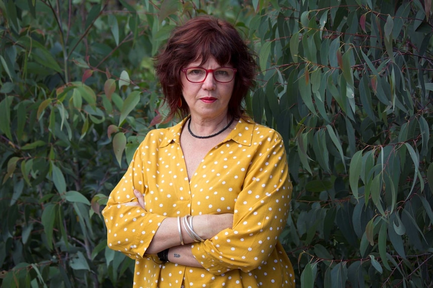 A woman stands of a gum tree wearing red glasses and a yellow shirt looking directly into the camera.