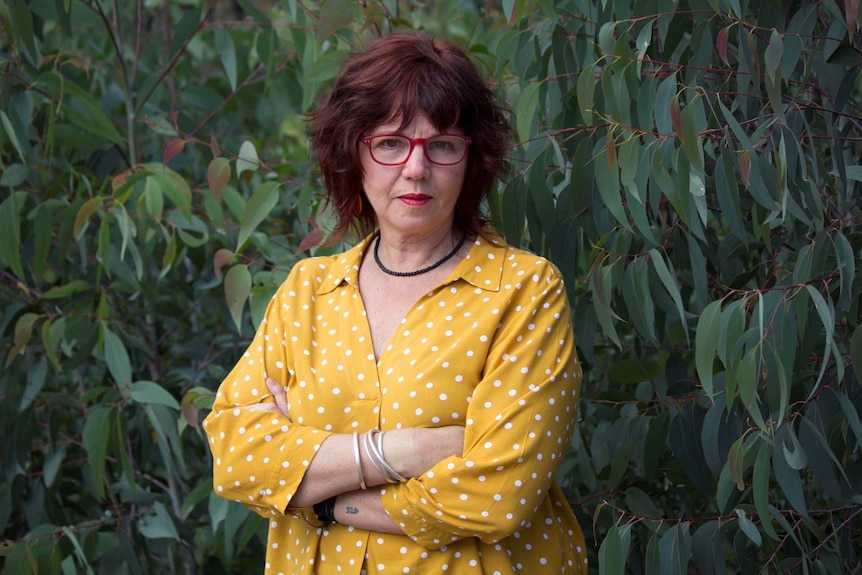 Jo Dodds stands of a gum tree wearing red glasses and a yellow shirt looking directly into the camera.