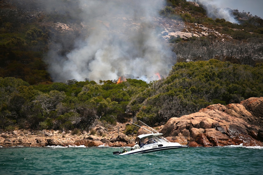 A blaze burns through scrub towards the ocean, as a man in a boat watches on.