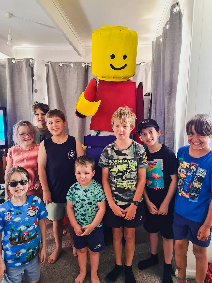 A tall man dressed in a lego suit stands next to a group of young children smiling.