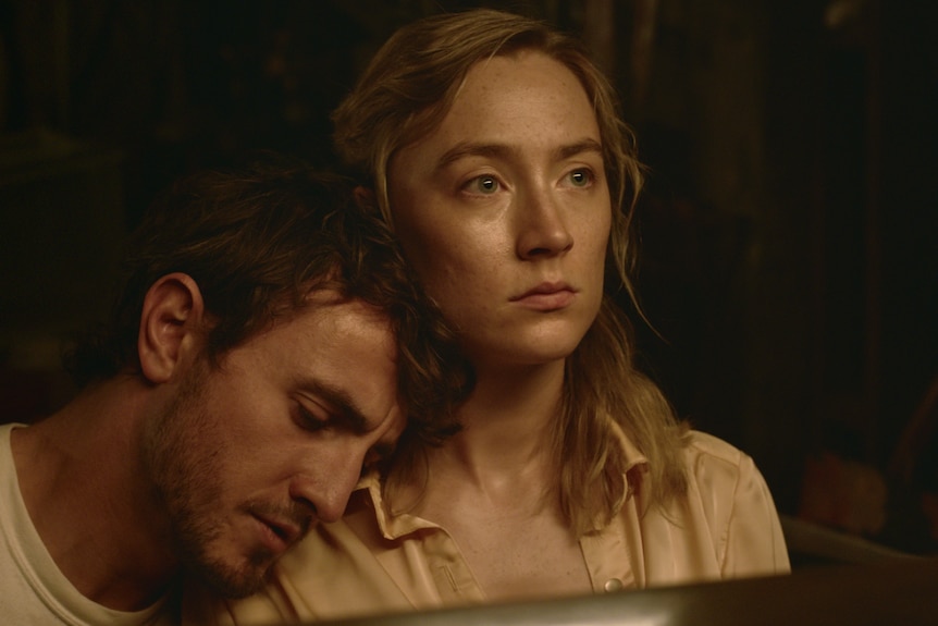 a young woman stares blankly ahead, a young man rests his head on her shoulder in a dark room.