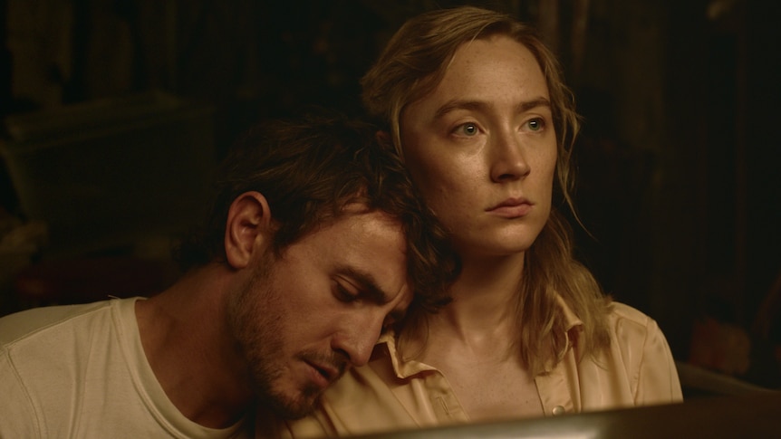 A white man leans his head on a white woman's shoulder while she looks pallidly in a bathroom mirror.