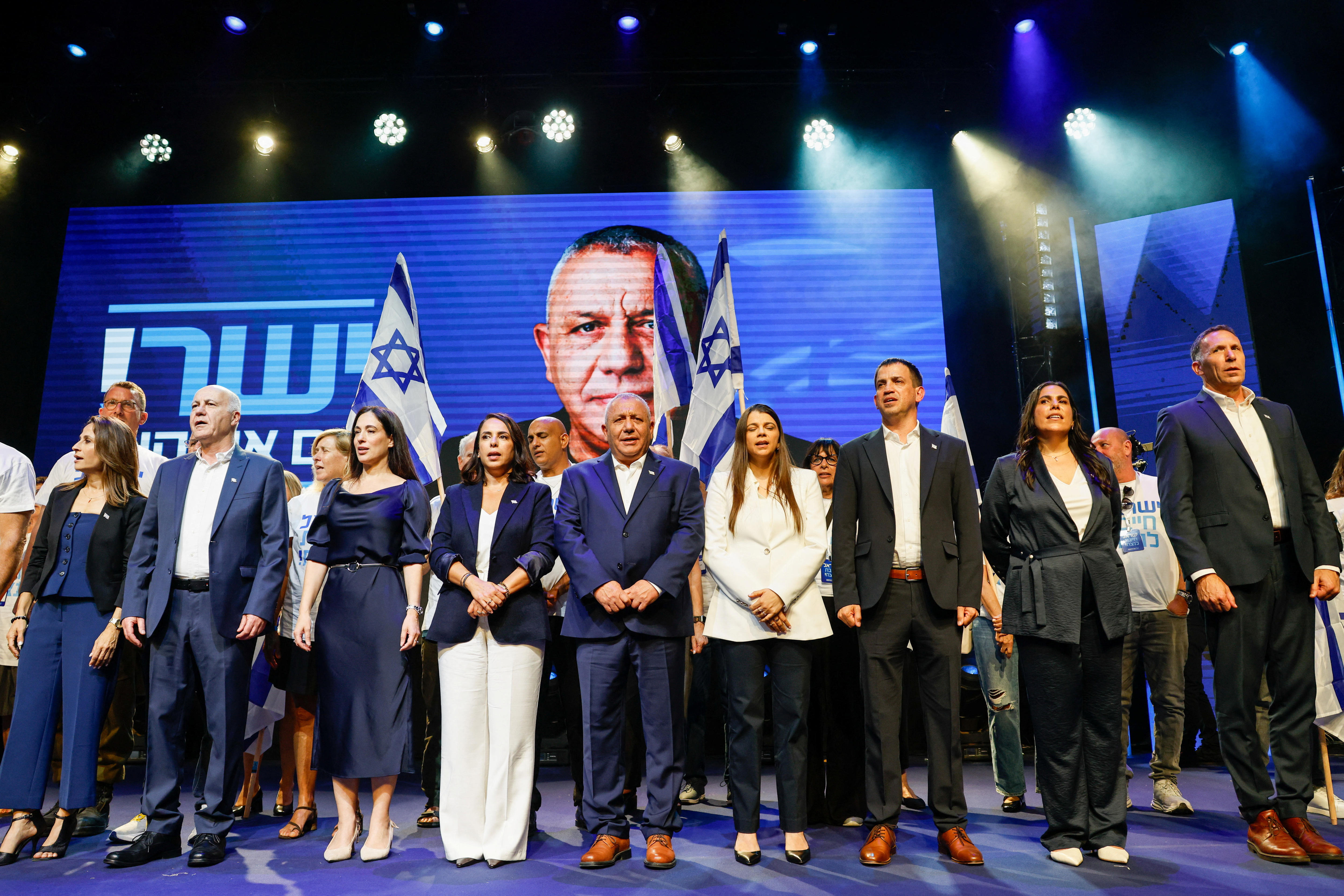 A line of men and women in suits and business attire signing in front of a large blue screen with the face of a man.