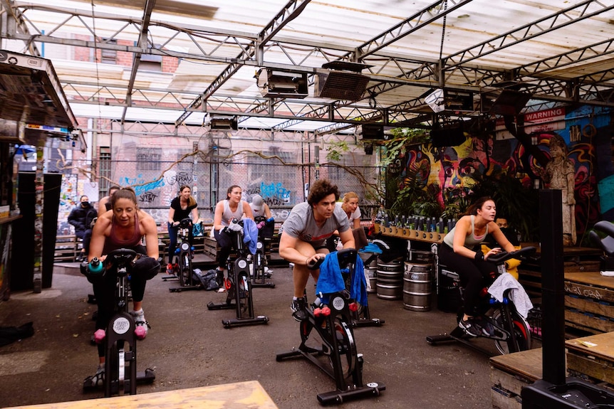 Seven people on indoor exercise bikes work out in a bar with graffiti walls.