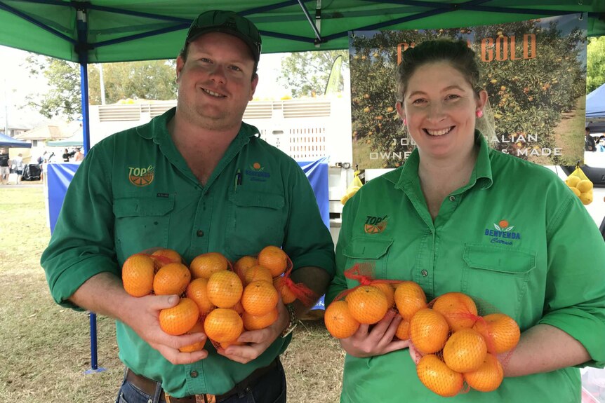 A man and a woman stand smiling at the camera, wearing green shirts, and holding bags of oranges.