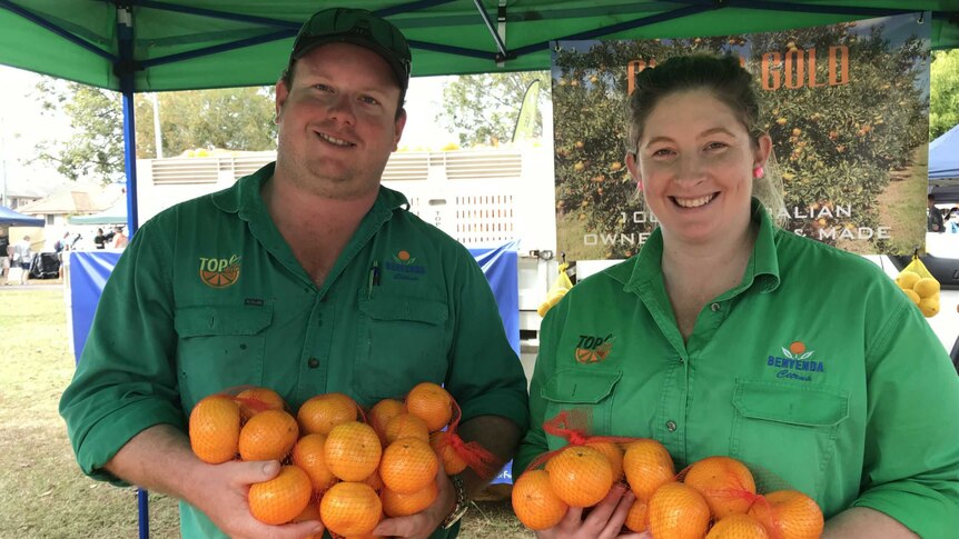 A man and a woman stand smiling at the camera, wearing green shirts, and holding bags of oranges.