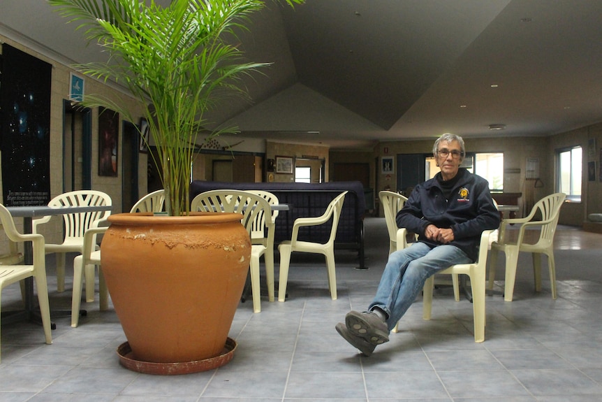 He sits in a plastic chair with his legs crossed looking at the camera with a giant pot plant next to him