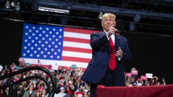 Donald Trump stands in front of a crowd and large flag at a rally, pointing