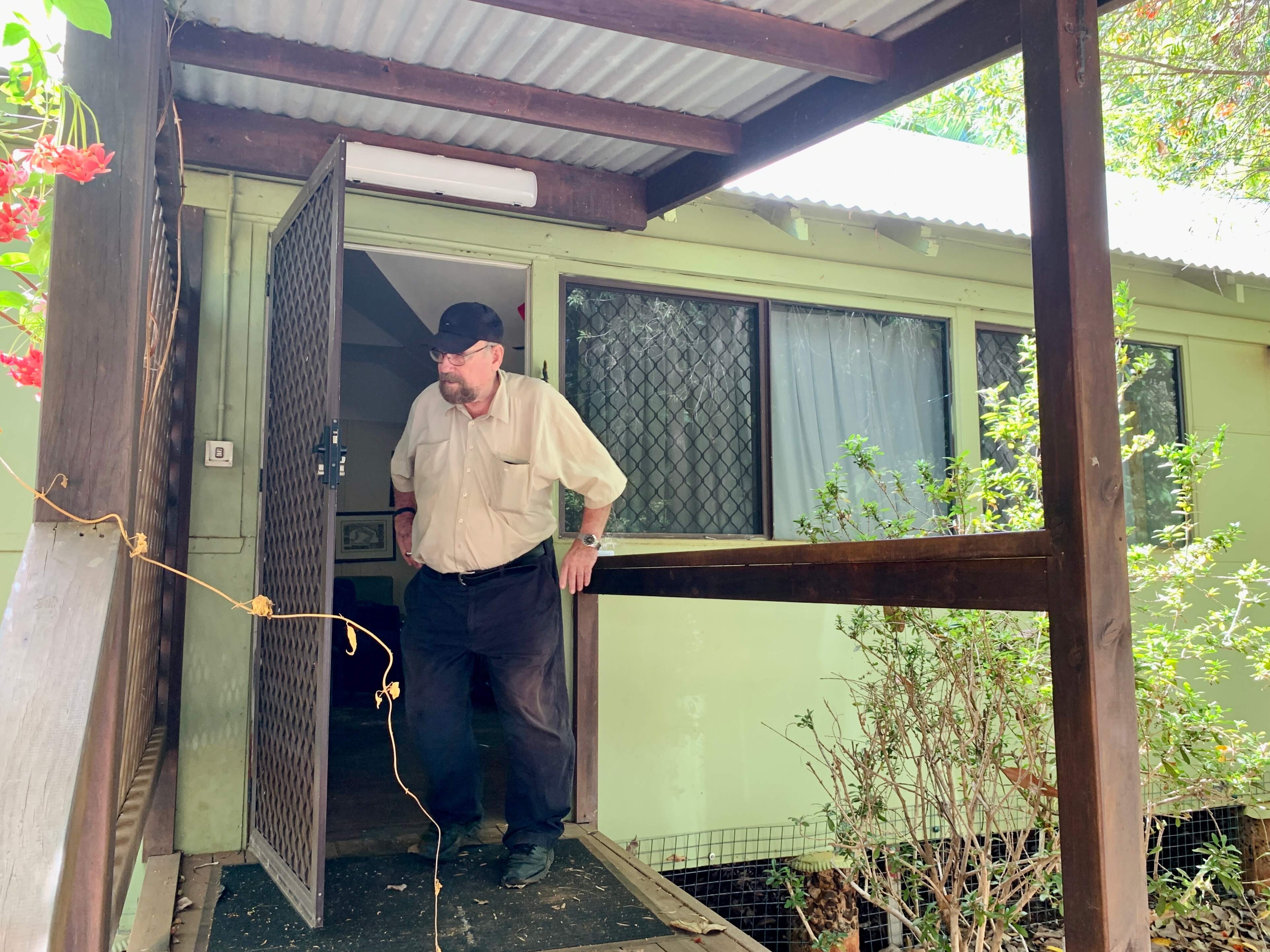 A man with a beard and wearing black cap stands partially turned from the camera in the doorway of a house.