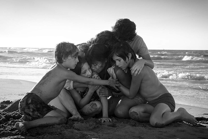 A black-and-white screen still shows six people huddled together on a beach. Waves crash gently behind them.