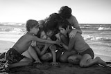 A black-and-white screen still shows six people huddled together on a beach. Waves crash gently behind them.