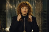 Woman with curly red hair standing facing camera as if looking at herself in mirror, holding her hair with each hand.